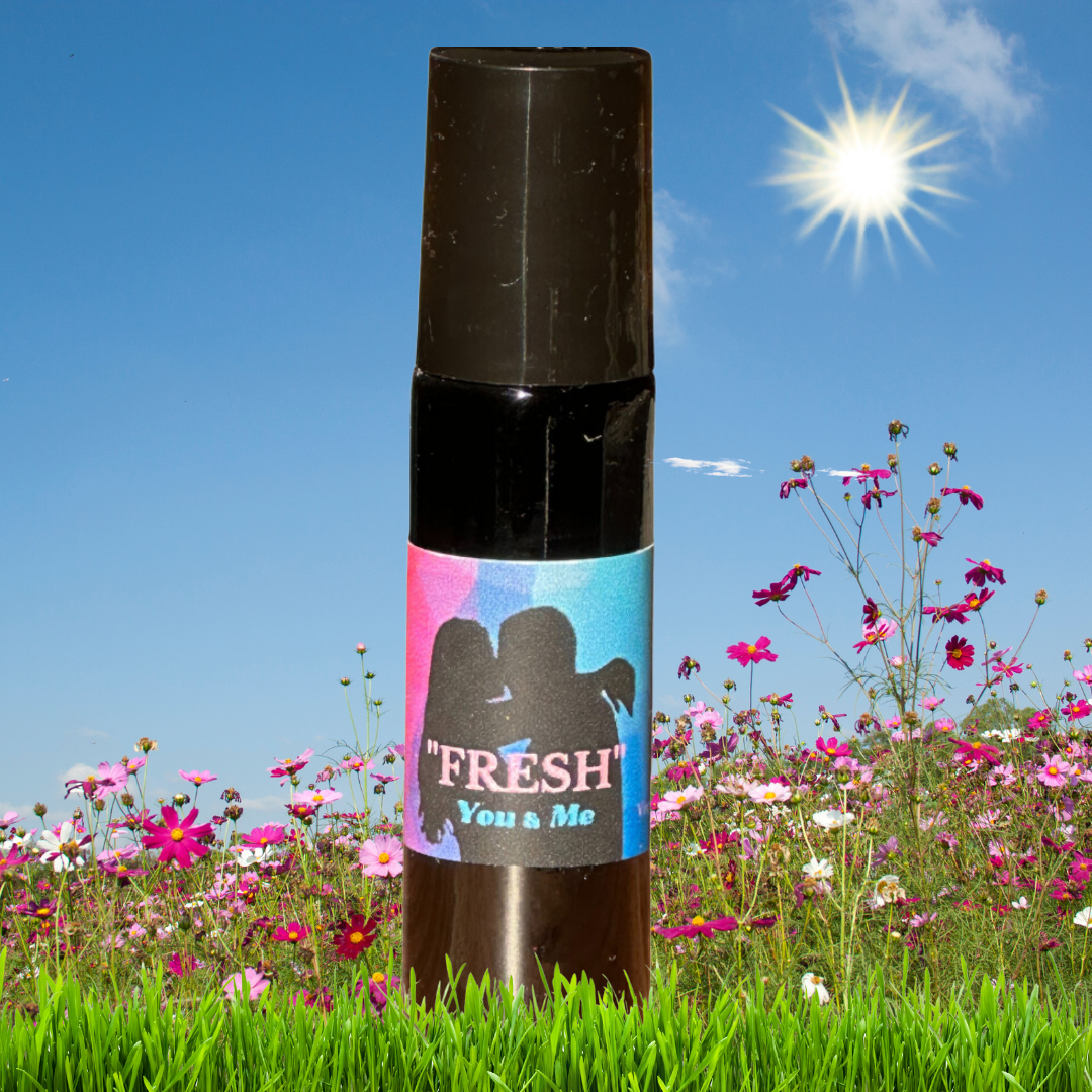 A 10 ml roll-on bottle of Viva Essensual Elixirs all natural, vegan perfume called Fresh, is in a field of flowers amongst the grass. The sun is shining high and bright in the blue summer sky. 