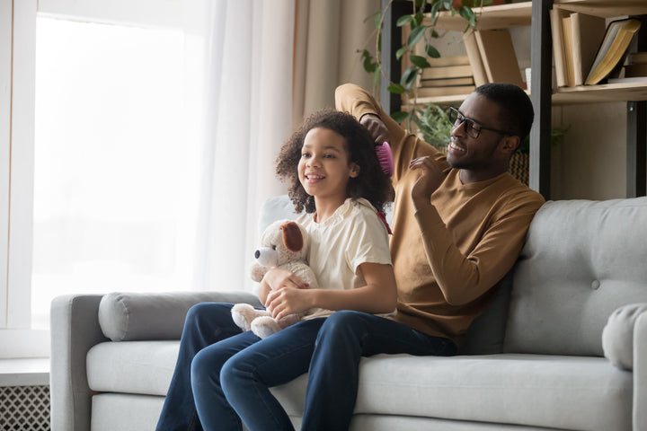 Handsome Black Dad sits on couch with beautiful Daughter gently combing her hair while she smiles and holds her stuffed toy dog. Bright light is shing into the room from the nearby window. A plant and books are behind the couch. Dad  wears glasses and a lovely smile enjoying this bonding time with his Daughter. A Dad who is gentle, nurturing & cares ... a Man of Integrity.