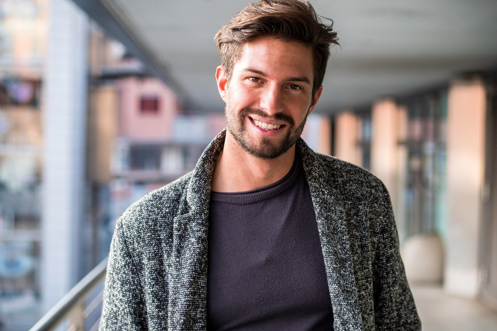 Richly intriguing without being too overpowering. His strength is in his character. Roll this 100% natural perfume on him or enhale and think of him. In this picture a handsome young European man smiles warmly into the camera. He's walking outside a university building wearing a Dark blue T shirt and grey mixed knit blazer. He's got well styled brown hair with layers on top, a moustache and beard with a little stubble. His head tilts to one side slightly as he looks at someone he will warmly greet.